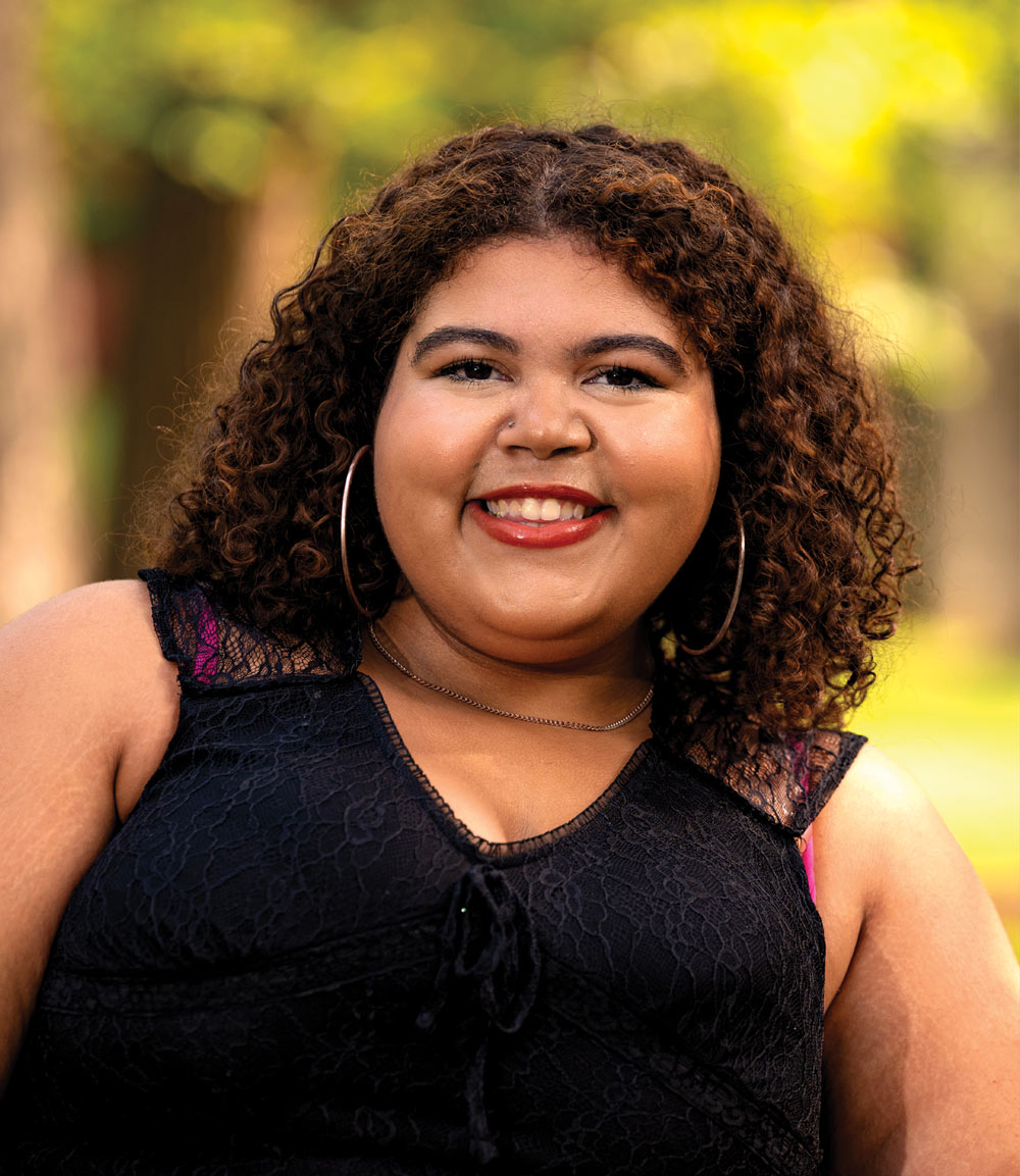 Smiling person with curly hair and a black lace top outdoors.