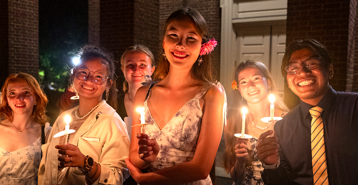 Group of young adults holding candles, smiling in evening attire with a brick backdrop.