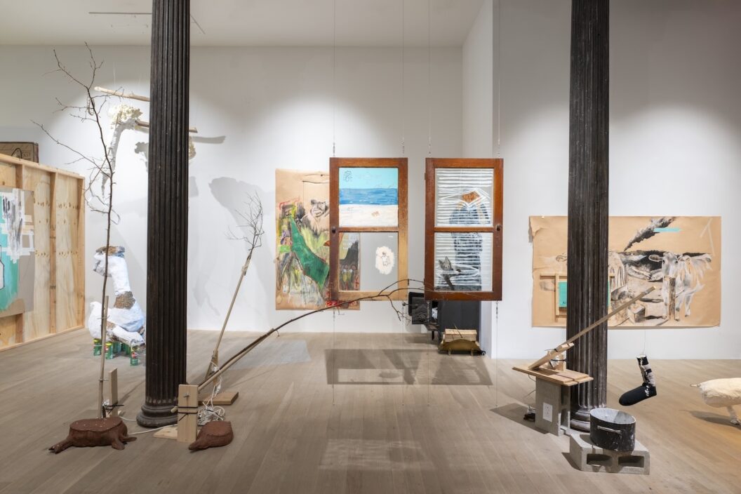 A photograph of a gallery installation with two large black ionic columns on either side of the image. Surrounding the left-hand pillar are sculpted appendages of trees, including stumps and branches, on the floor screwed to different pieces of wood. Behind the column sits a paper maché sculpture sitting atop cans of Comet bleach. To the left and right of the sculpture are two paintings on cardboard and brown paper. In the center of the gallery space are two wooden window frames hanging from the ceiling. The glass of the windows are painted on, with the left frame depicting a seascape and the right frame  a figure and a cat peeking through blinds. In front of the right-hand column there is a sculpture consisting of two concrete blocks. Atop one block are two wood planks where a wooden rod sits above them at a 45-degree angle. A sock, tied to a wire hanging from the tallest end of the rod, dangles above a large cooking point whose interior has been painted in with white. The pot also sits on a concrete block. Behind the right-hand column is a painting on brown paper depicting a cow and a landscape with a cloud of smoke in the distance.