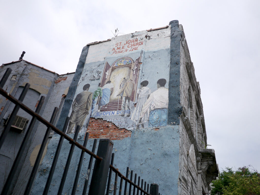 A photograph of a mural. The wall is coated in cement, which is falling away to expose the red brick beneath. In the mural, which has muted colors, people are lined up on either side of an aisle to walk through doors adorned with crosses and enter into a light-filled space. Above the doorway is “St. John A.M.E. Church. Praise the Lord.