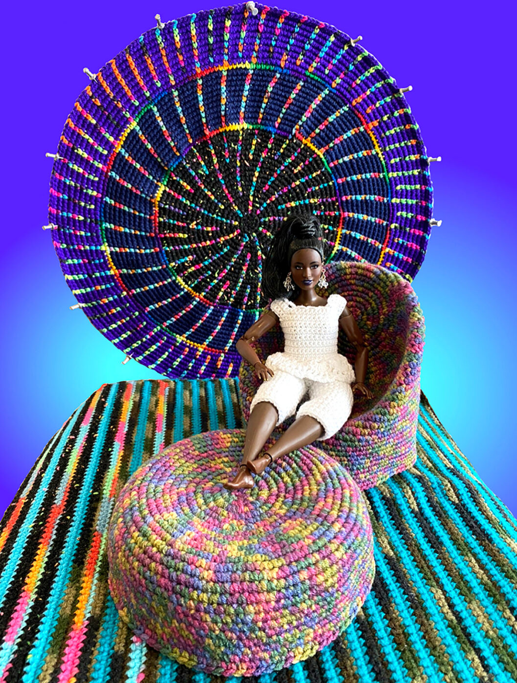 1212 A doll wearing a white dress is seated in a crocheted chair with a matching crochet ottoman. The furniture prototypes are placed on a crocheted rug that is teal, black, yellow, and run. Behind it is a circular crochet form, with purple, pink, yellow, and blue yarn.