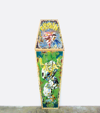 A coffin-shaped canvas with yellow edges on a gray floor and resting against a white wall. The painting features countless turkey hands in blue and green superimposed upon one another all throughout, as well two instances of the word “organ” written in yellow capital letters.