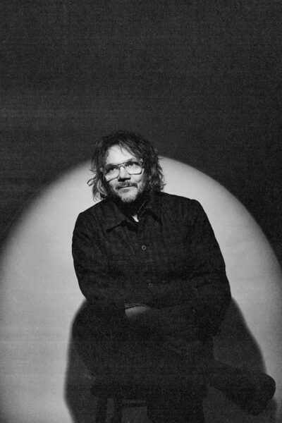 A black-and-white portrait photograph of a man with shaggy hair and glasses sitting on a stool. He is spotlit and has one leg crossed over his knee. He has a faint smile.
