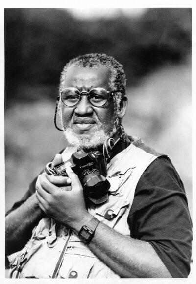 A black and white photograph of Shawn Walker. He is wearing a t-shirt and fishing vest; clutching a Canon camera to his chest and squinting through his glasses. He appears middle aged, with an unkempt beard.