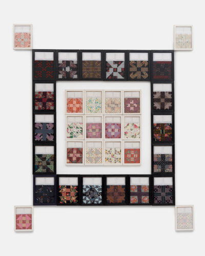 Eighteen black-framed panels surround a four-by-three grid of white-framed panels. Four more white-framed panels extend from the corners of the surrounding panels. Each panel shows a unique quilt with two small pillows. The assemblage is hung on a white wall.