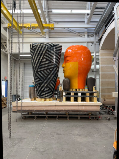 A tall oblong ceramic work with black stripes and a large ceramic orange head appear on a platform in an industrial space. The platform also supports smaller oblong ceramic works.