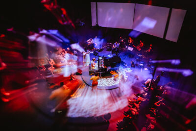 Samora Pinderhughes playing piano while surrounded by an accompanying band on a circular stage. The lights cast on the room are blue, white, and red. An audience sat in chairs circles the musical troupe.
