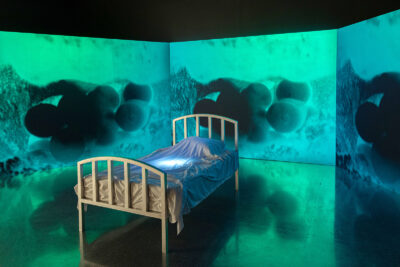 A photograph of a single bed surrounded by a three-wall, turquoise projection of an obscured image of what appears to be eggs.