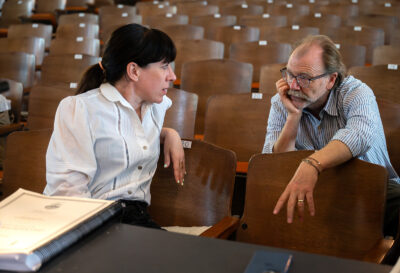 Missy Mazzoli, in a white button-up shirt, sitting in a theater seat and looking back at George Saunders, in a striped button-up shirt, sitting in the row behind her.
