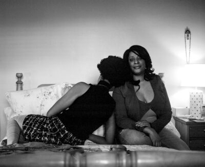 A black-and-white photograph of two people sitting on a bed. The figure on the left is turned away from the camera and leaning on the other person’s shoulder.
