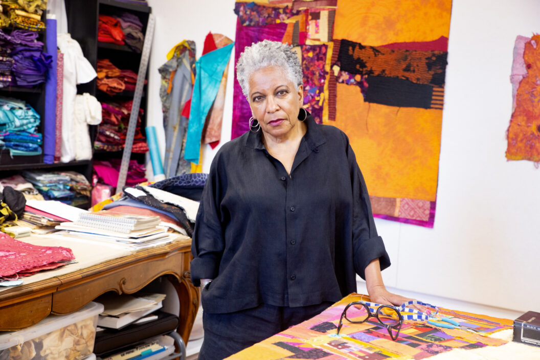 Harris, a Black woman with gray hair and pink lipstick, stands in her studio surrounded by textiles in vibrant colors.