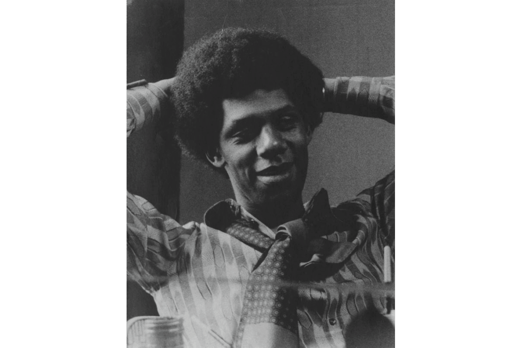 A black-and-white photograph of Peter Bradley, a Black man with an afro, in a printed shirt and tie, reclining with his arms behind his head.