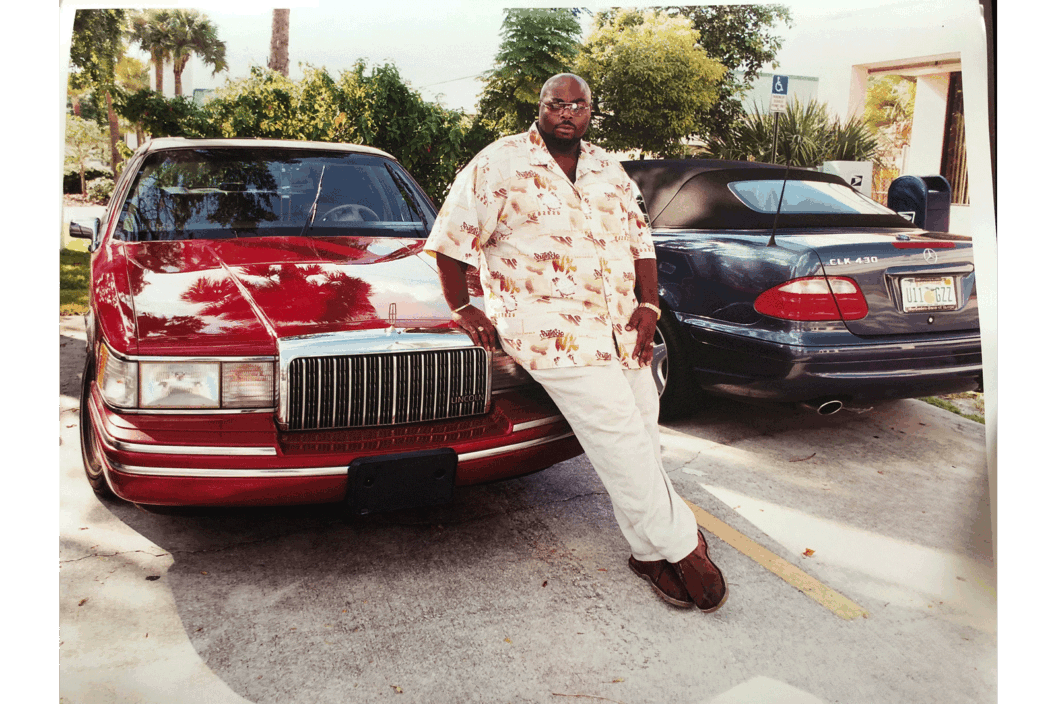 Color photograph of a man leaning against a red car.