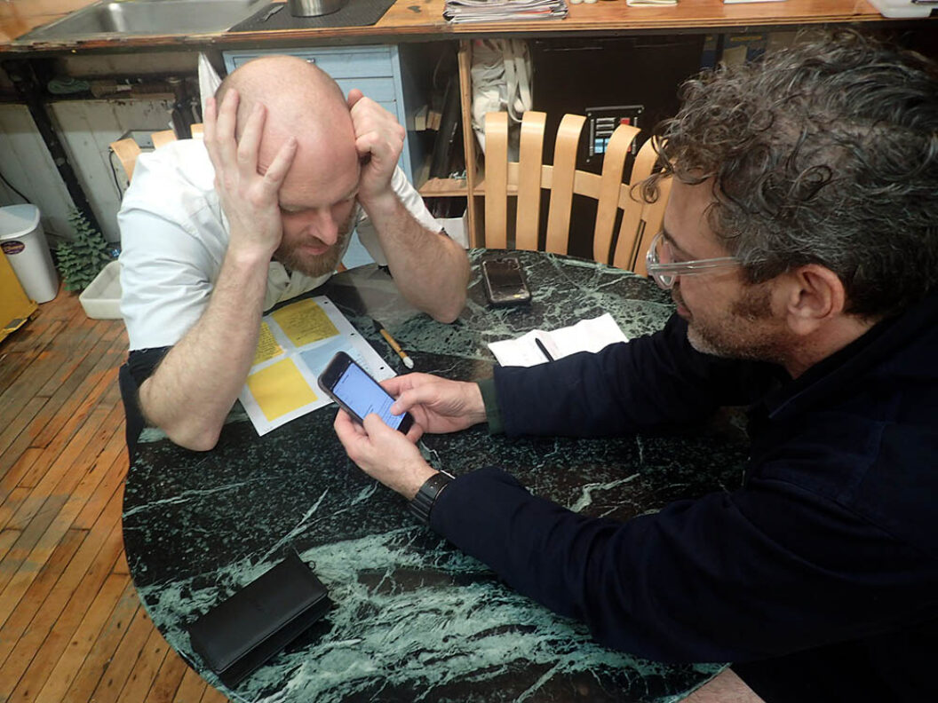 Filmmaker Van Neistat sits at a round marble table with Tom Sachs. Neistat is bald, wearing a white shirt, and has his head in his hands. Across from him, Sachs wears a black shirt and clear rimmed glasses, and is scrolling on his phone. On the table are loose leaf papers, pens, and a phone. Behind the table is a studio space with hardwood floors.
