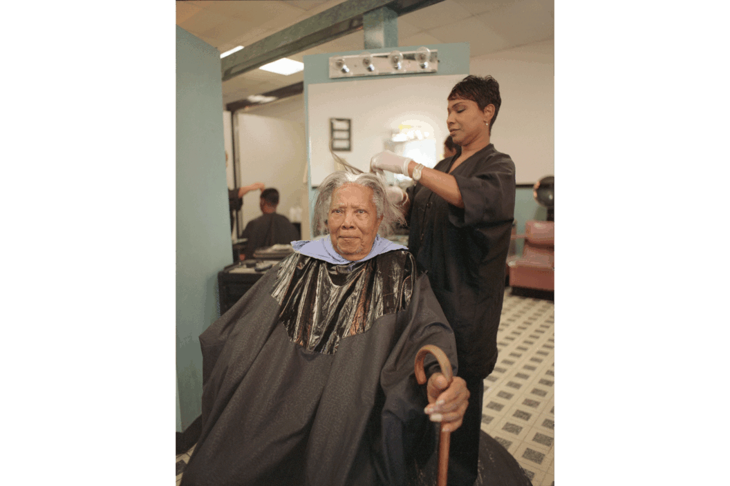 Color photograph of a woman holding a cane as another woman does her gray hair.