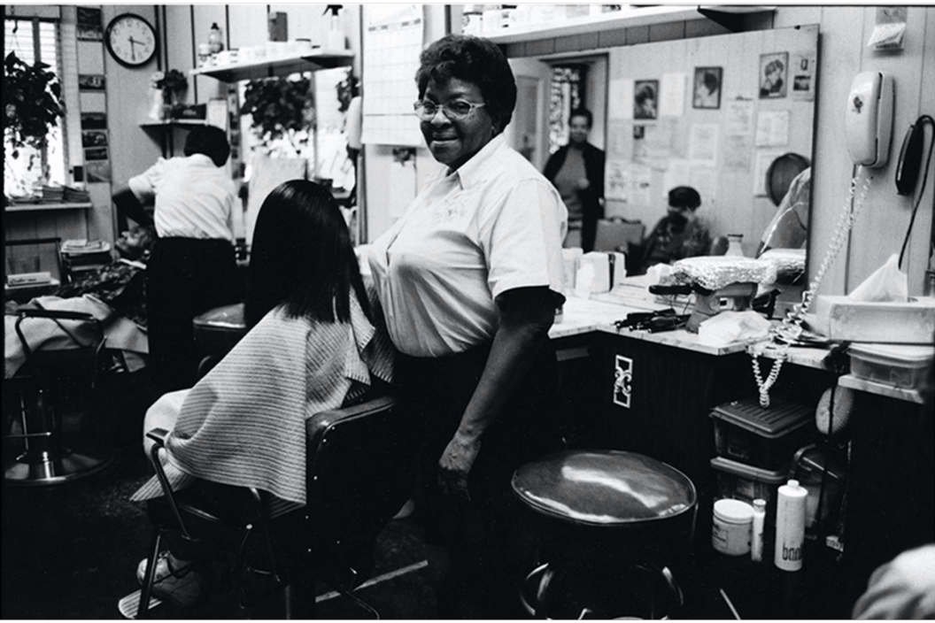 A woman smiling at the camera, looking like she is styling the hair of the person in the chair in a beauty salon.