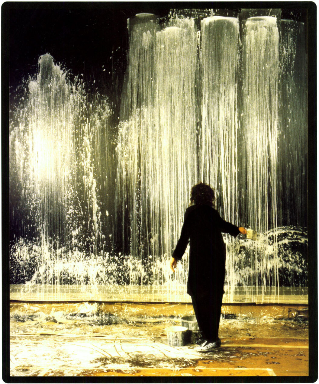 A color photograph of a woman with dark hair wearing dark, loose clothing throwing light paint on a dark surface. She stands in a large space with painting tools and paints around her feet, on the floor.