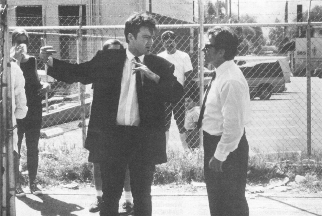1400 A black-and-white photograph of two male-presenting people conversing in front of a chainlink fence while people watch from in front of and behind the fence.