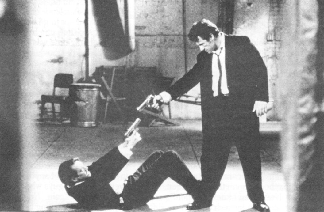1400 A black-and-white photograph of two male-presenting people, both dressed in suits, pointing handguns at each other. The person on the left is on the ground and the person on the right is leaning over the other.