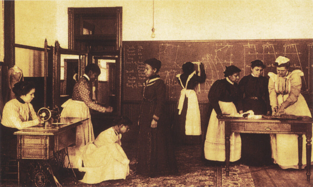 A room with drawings is filled with multiple female-presenting people. Many have dark skin and two have light skin and all are dressed in full-length dresses. All are intensely focused.