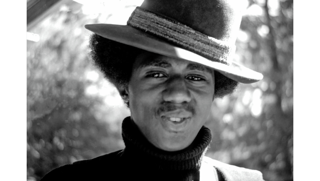 A black and white photograph of James LIttle in 1971. He is wearing a turtleneck and a fedora, and is in the middle of speaking.