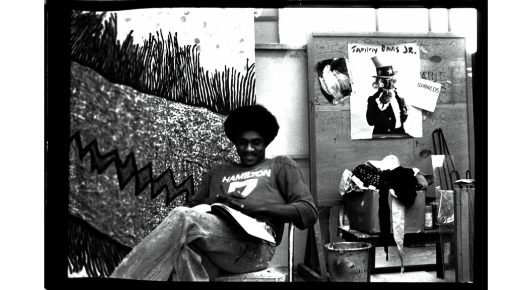 A black and white photograph of James Little at his studio in Memphis as he sits relaxed in a folding chair and smiles while working from a sketchbook.