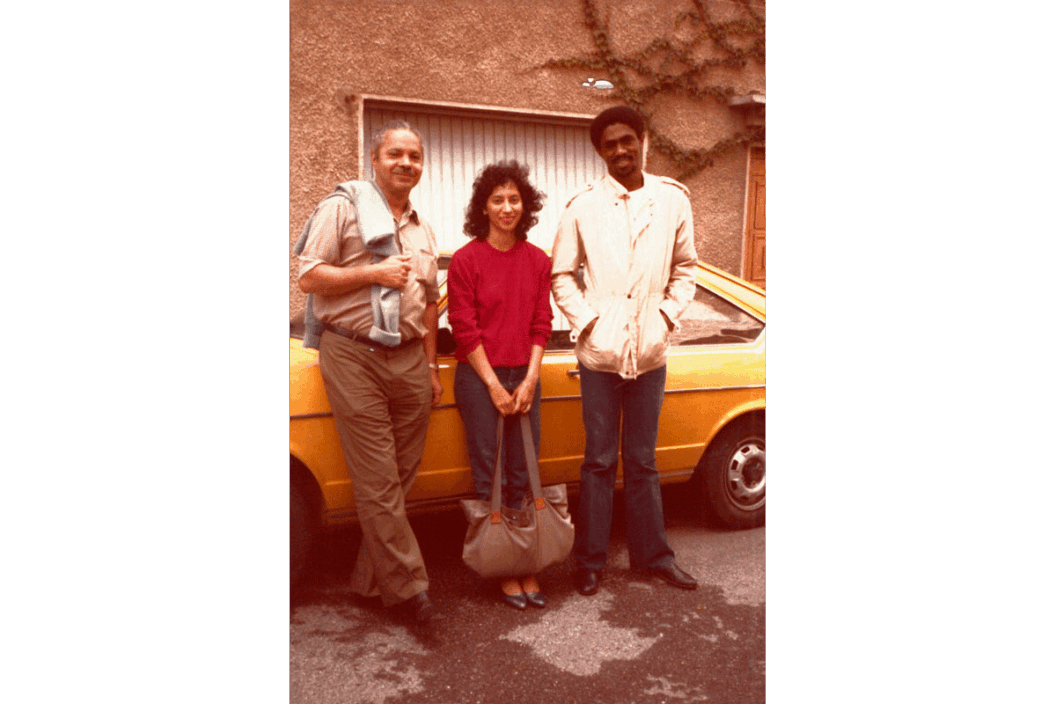 A photo of Harold Hart, Fatima Shaik, and James Little standing in front of a yellow car. The three artists are standing outside of a brown stucco building on rainy asphalt, and dressed in light jackets and jeans.