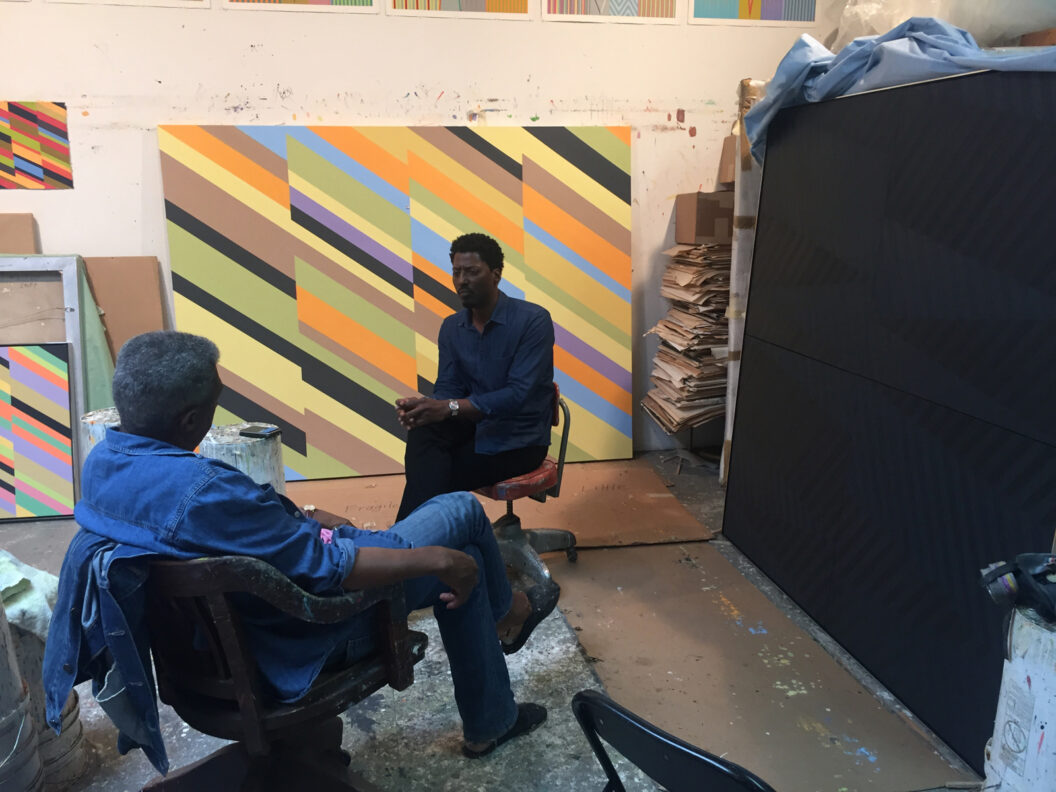 A 2016 photograph of James Little and LeRonn P Brooks sitting in chairs while chatting in front of large canvases at Little's studio in Williamsburg, Brooklyn.