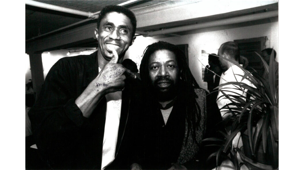A black and white photograph of James Little and Ellsworth Ausby from 1996. Little is posing with a thumbs-up gesture while Asuby looks into the camera.