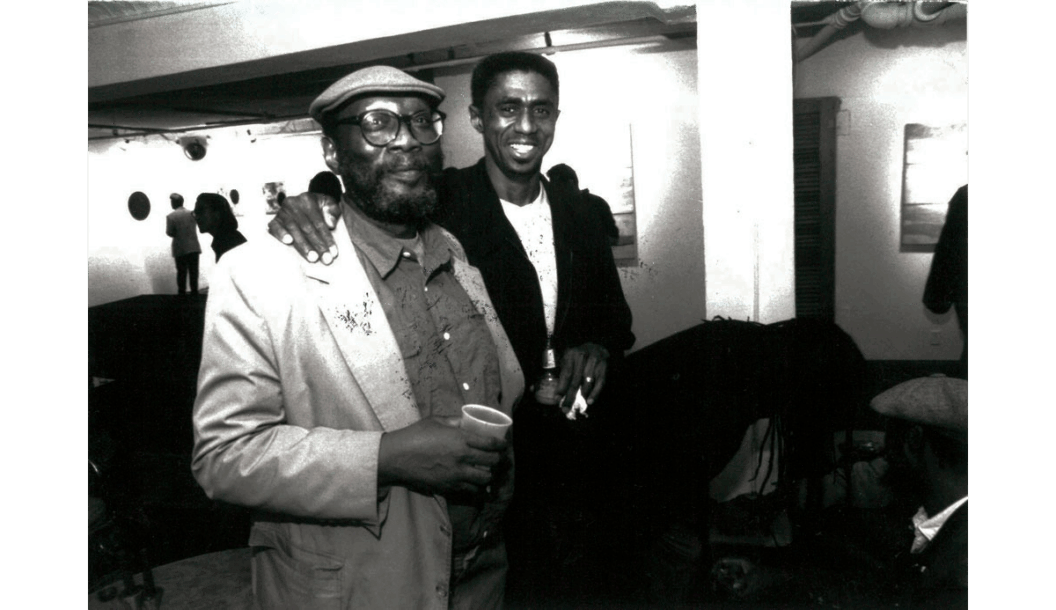 A black and white photograph of James Little with his arm around Frank Bowling at a gallery in 1996.