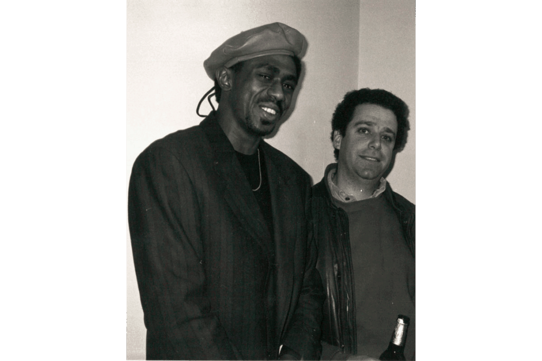 A black and white photo of Little and Ken Perkins at Kenkeleba Gallery. Little is wearing a black shirt with a blazer and a chain necklace with a leather beret on his head. Perkins wears a crewneck sweater over a collared shirt, and he is holding a bottle of alcohol.