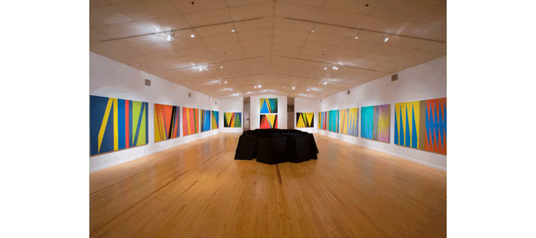 Wide-angle shot of Little's paintings on display at the Station Museum of Contemporary Art, along with a sculpture by George Smith on the floor. The sculpture is a cluster of deep black cubes and contrasts with Little's bright and multi-color paintings.