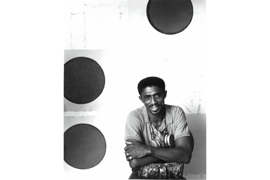 A picture of James Little, a Black man, in his Chelsea art studio. He is folding his arms and smiling while leaning on the back of a chair.