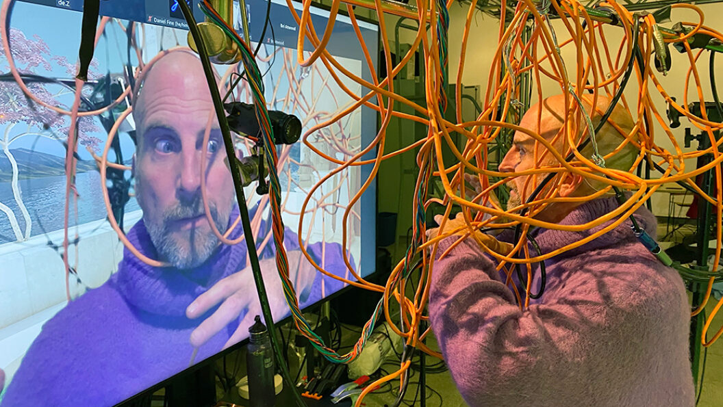 Man engulfed in orange wires, facing a tv screen projection of himself.