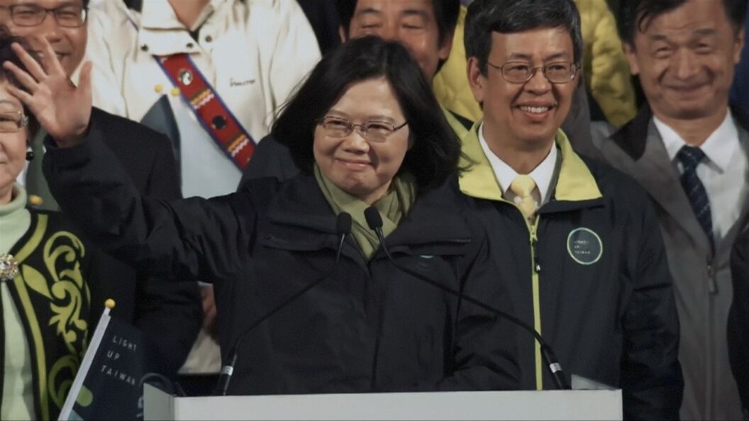 President Tsai, a middle-aged Taiwanese woman with glasses and shoulder-length black hair, waves to a crowd beyond her podium. Behind her, a large group of government officials in formal dress and winter-wear smile.