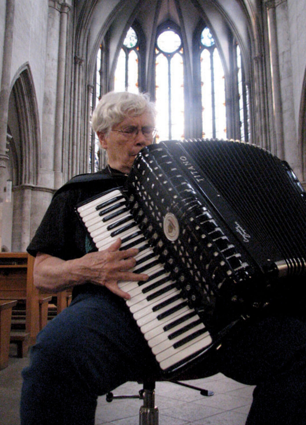 Photograph of Oliveros, white man, sitting on a stool playing an accordian in a church.