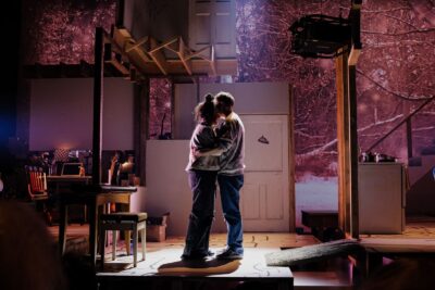 A husband and wife embrace at the center of a dim spotlight. The stage around them is decorated like a cluttered house, the projections on the walls resembling a galaxy.