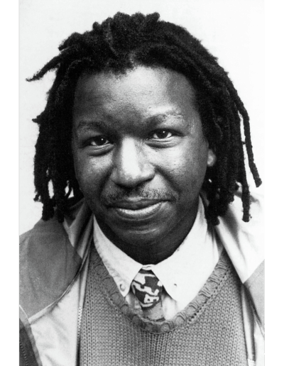 A black and white studio photograph of a Black man with mid-length locs, smiling at the camera. He wears a tie, white collar shirt and a sweater vest while shown on a white background.