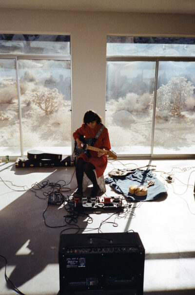 A woman sitting in a room playing a guitar with a desert landscape behind her.
