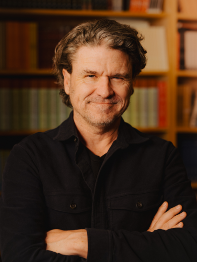 A portrait photo of Dave Eggers looking at the camera, smiling, and crossing his arms.