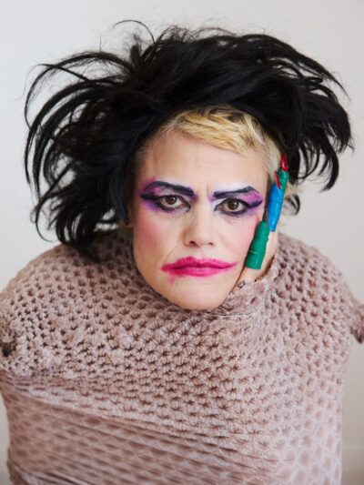 A headshot of Dynasty Handbag in a beige mesh sack with smeared clown-color goth makeup and a ratty blond-black Siouxsie-Sioux wig.