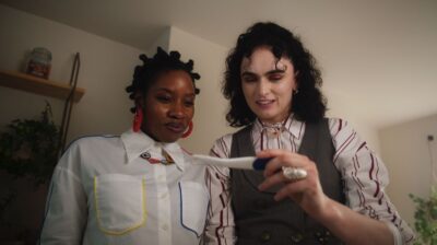 The couple, both dressed in button-down shirts with accessories, check a pregnancy test in their dim bathroom.