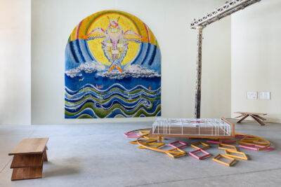 An installation view of a large tapestry, a sculptural bed, and two wooden benches.