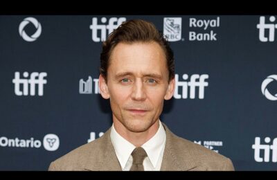 A closeup press photo of British actor Tom Hiddleston against a TIFF-branded backdrop. He is pale with short brown hair and a tan suit.