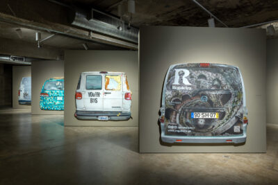 An installation view of paintings of the back of four vans on separated, staggered walls.