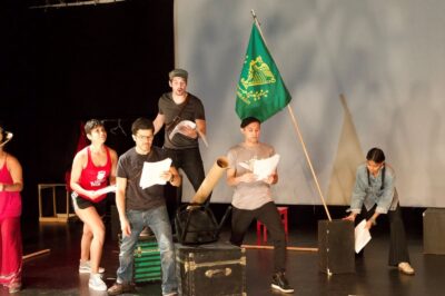 Performers in rehearsal clothes, scripts in hand, stand in a diamond formation with the upstage actor atop black boxes. To their right, another actor raises a green flag.