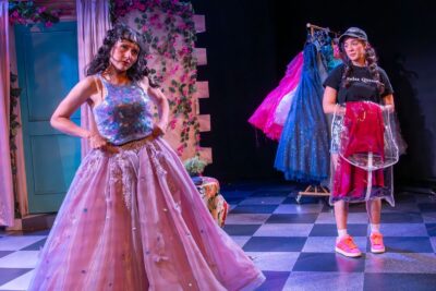 A girl tries on a two-piece dress at the front of the stage. The top is a purple patterned tank and the bottom is a pink tulle skirt, and her friend waits by the dress rack with a red dress in a garment bag.