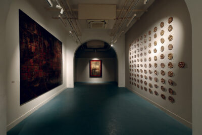 An installation view of small metal sculptures installed on a wall across from a large abstract painting in squares and a shiny abstract painting through a doorway.