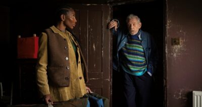 The painter and assistant confront each other in a dark brown hallway. The painter is leaning against a doorframe in a striped blue sweater while the assistant, in profile, wears layered earth tones.
