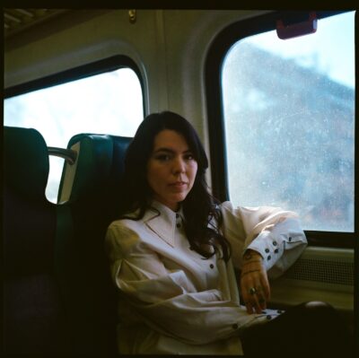 T Kira Māhealani Madden sits in a train seat, wearing a white button-up shirt, arm resting on the window ledge, looking directly at the camera.